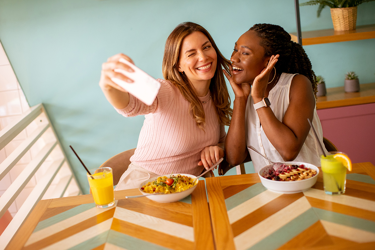 Two pretty young women, caucasian and black one, taking selfie with mobile phone in the cafe